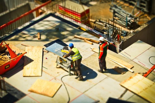 Two construction workers on a building site operate a table saw, surrounded by plywood sheets and construction materials. Both wear safety gear including hard hats and high-visibility vests. The site is partially enclosed by barriers.