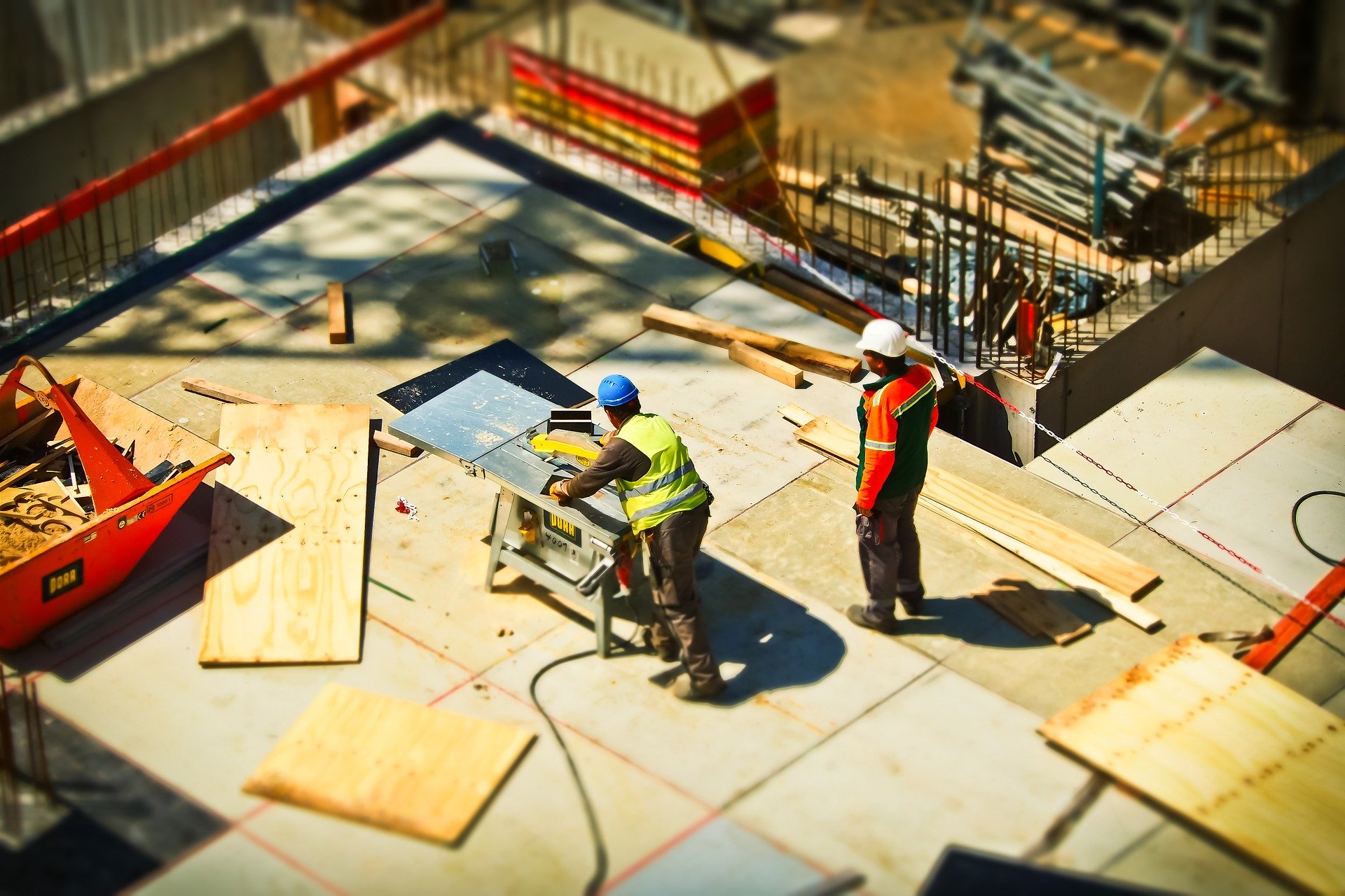 Two construction workers on a building site operate a table saw, surrounded by plywood sheets and construction materials. Both wear safety gear including hard hats and high-visibility vests. The site is partially enclosed by barriers.