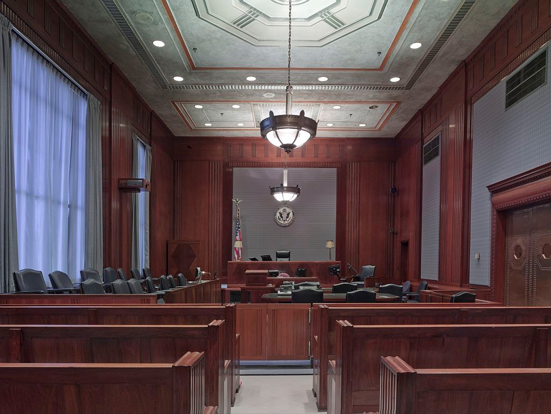 Image of an empty courtroom with wooden walls and furnishings. Rows of benches are visible in the foreground, leading to a judge's bench and witness stand. An American flag and a seal are displayed behind the judge's area. Chandeliers hang from the ceiling.