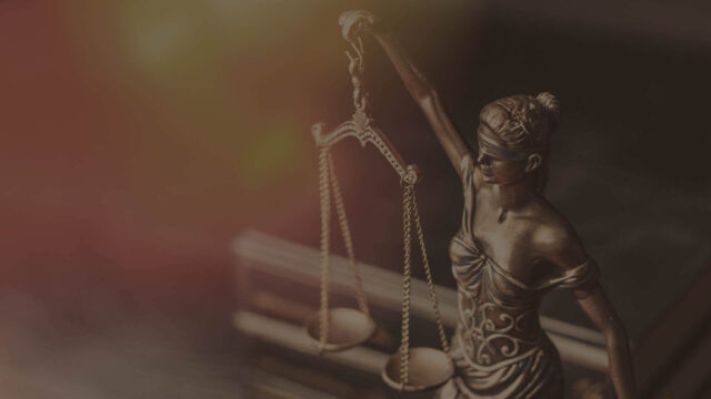 Close-up of Lady Justice statue, holding scales with a blindfold covering her eyes, symbolizing fairness and impartiality. The background is softly blurred with warm colors.