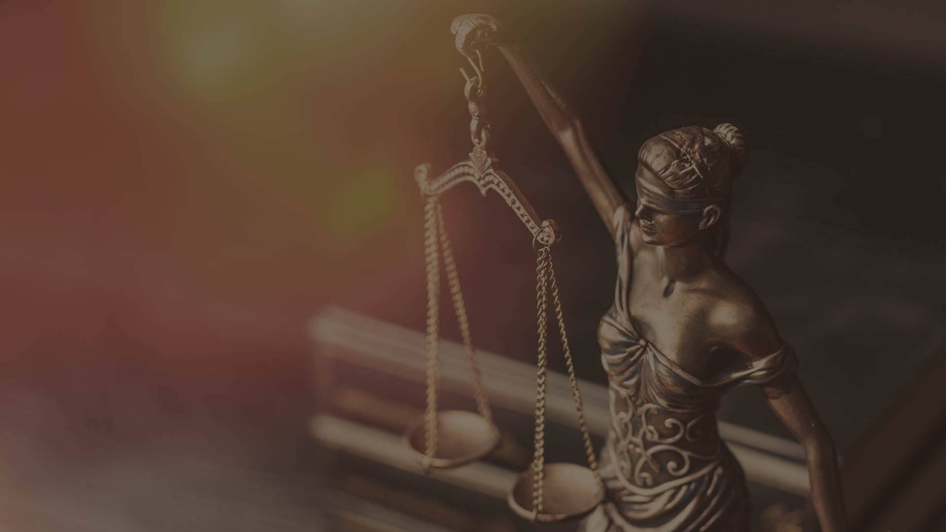 Close-up of Lady Justice statue, holding scales with a blindfold covering her eyes, symbolizing fairness and impartiality. The background is softly blurred with warm colors.