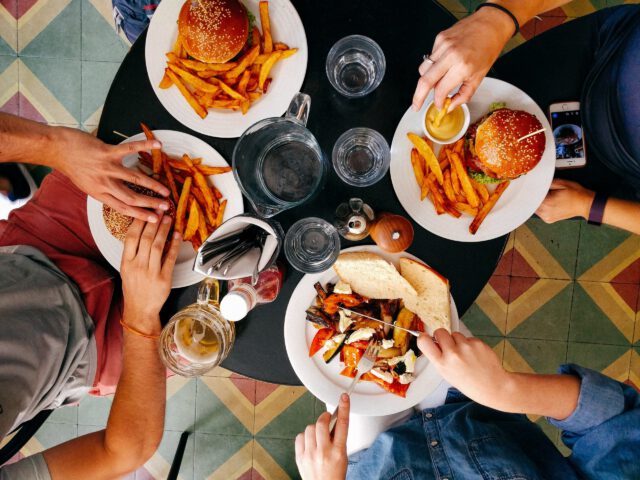 A top-down view of four people enjoying a meal at a Texas restaurant. There are two burgers with fries, a salad, and drinks on the table alongside utensils and a smartphone. Colorful geometric tiles add to the ambiance, showcasing what to know about dining in style.