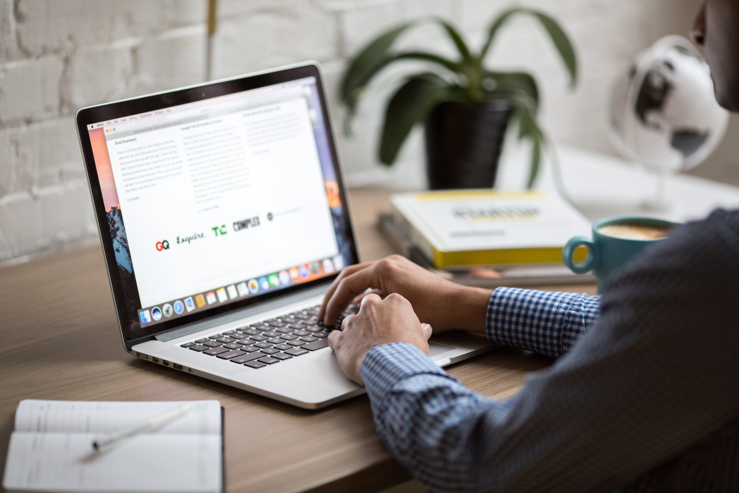 A person in a checkered shirt is typing on a MacBook laptop at a wooden desk, researching identity theft penalties in Texas. A potted plant and books are in the background, while the screen displays an article with text and logos. A mug sits nearby.