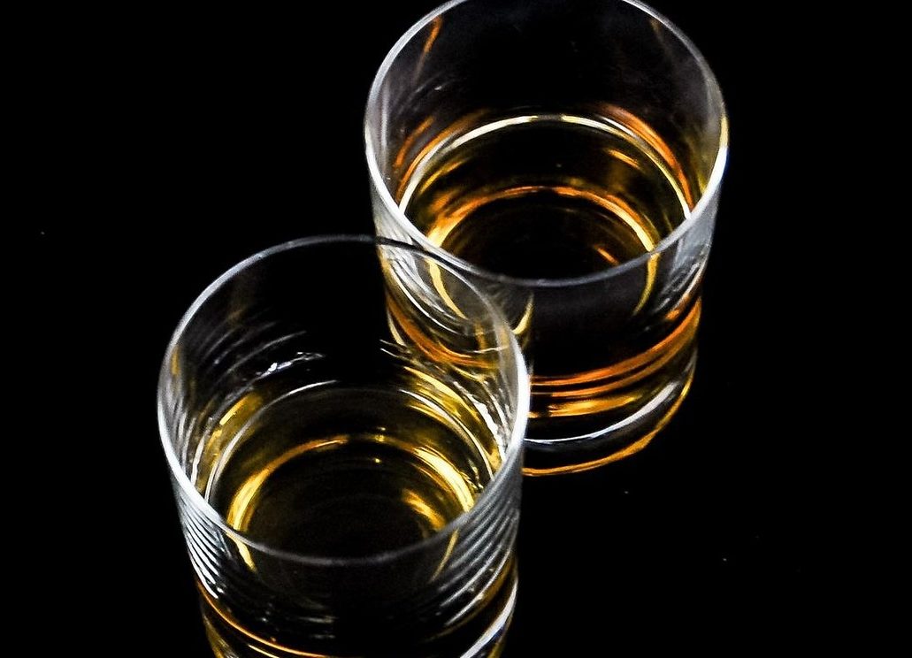 Two clear glass tumblers filled with a golden liquid, possibly whiskey, stand against a black background. The lighting casts reflections on the glasses, highlighting the rich color reminiscent of a Texas sunset.