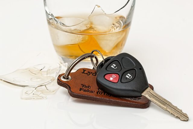 A car key with a black remote rests on a brown leather keychain beside broken glass and a half-filled glass of amber liquid on a white surface.