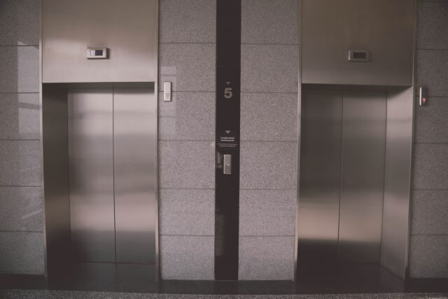 A modern elevator lobby with two closed elevator doors. The walls are made of sleek, gray stone. An indicator above each door shows the elevator's status. The number 5 is displayed on the wall between the elevators.