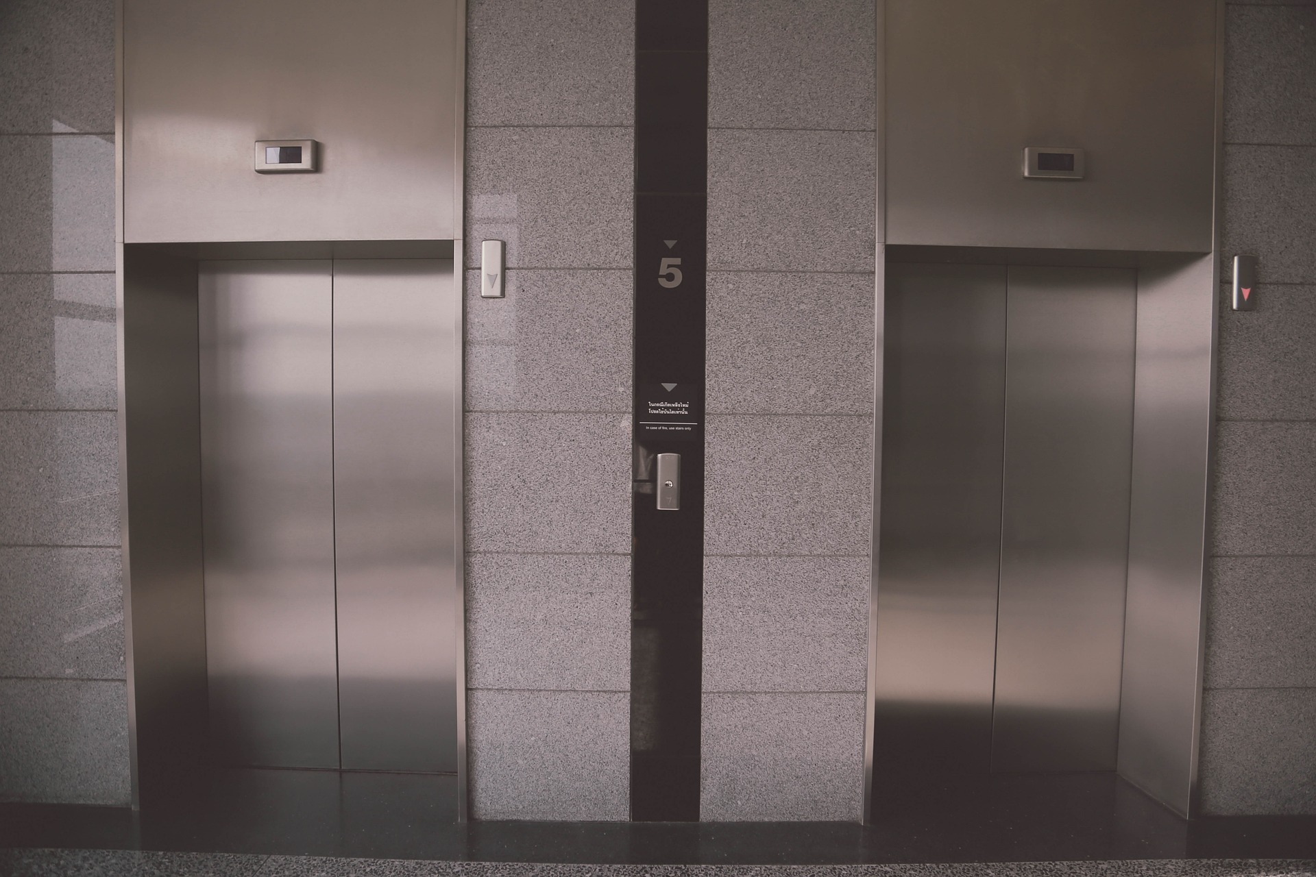 A modern elevator lobby with two closed elevator doors. The walls are made of sleek, gray stone. An indicator above each door shows the elevator's status. The number 5 is displayed on the wall between the elevators.