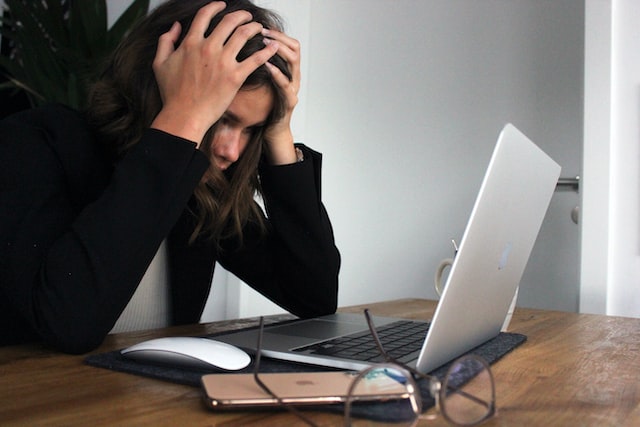 A person sits at a desk in frustration, their head in their hands while staring at a laptop screen. Glasses, a smartphone, and a computer mouse are scattered across the desk. In Texas, where cyberbullying can be considered a criminal offense, this scene is all too familiar.