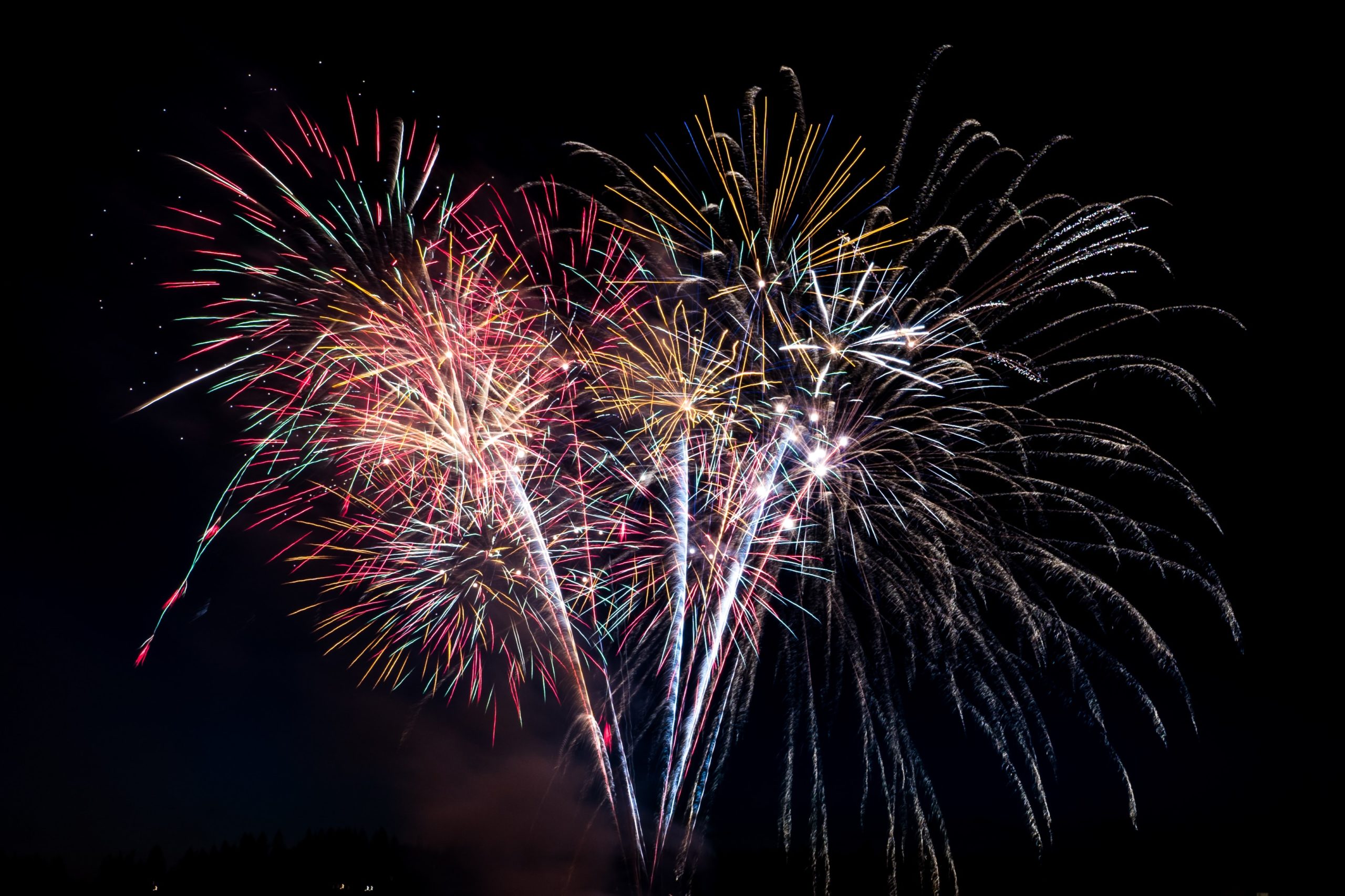 A vibrant fireworks display lights up the Fourth of July night sky with bursts of red, yellow, blue, and green against a dark background. Multiple fireworks explode, creating a dazzling and colorful spectacle while firework safety remains top of mind for all attendees.