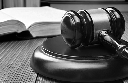 A black and white image of a judge's gavel resting on a sound block in the foreground, with an open book in soft focus in the background. The scene conveys a sense of law and justice.