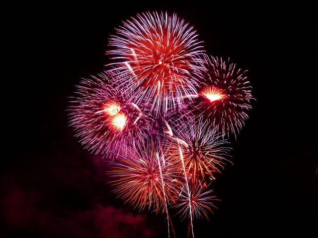 A vibrant display of fireworks in various colors, including red, pink, and purple, bursts against a black night sky. The bright patterns create a festive and celebratory atmosphere.