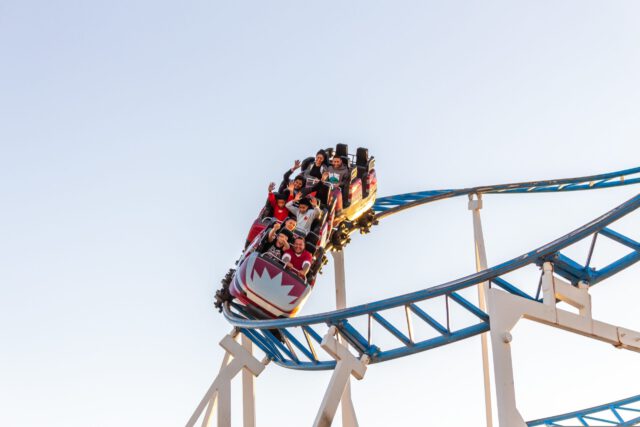 A group of people ride a roller coaster with their hands up, showing excitement and thrill at a Texas theme park. The coaster races along a blue and white track against a clear sky background.