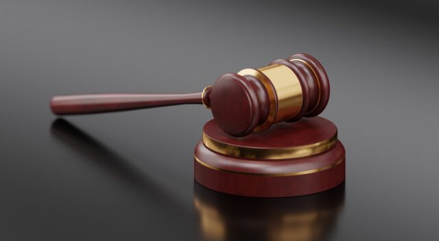 A wooden gavel with a gold band rests on a matching round sound block, set against a dark, reflective surface. The gavel and block are polished, with rich wooden tones highlighting the craftsmanship.
