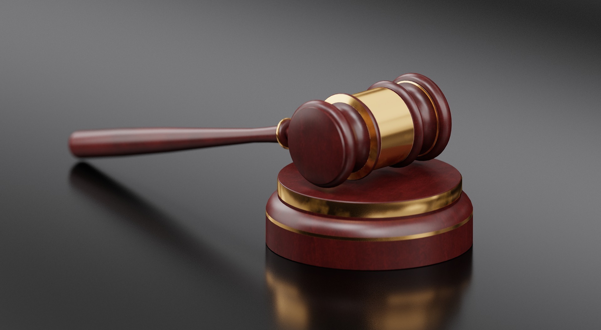 A wooden gavel with a gold band rests on a matching round sound block, set against a dark, reflective surface. The gavel and block are polished, with rich wooden tones highlighting the craftsmanship.