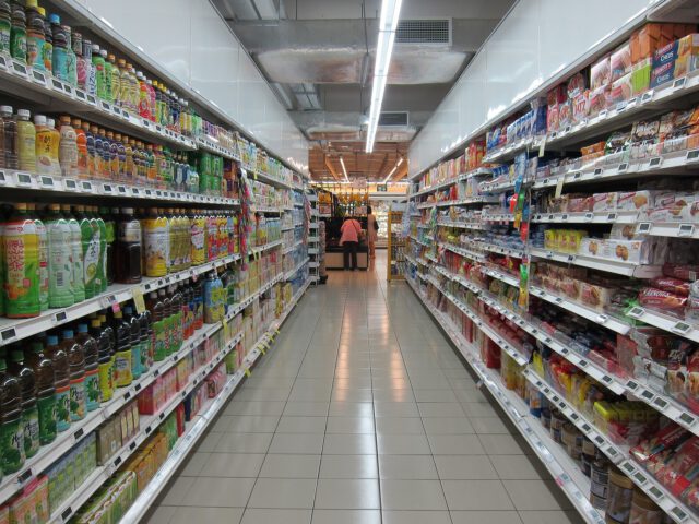 A grocery store aisle in Texas features shelves filled with various beverages on the left and packaged goods on the right. The tiled floor gleams under fluorescent lights, casting a charged glow. At the end of the aisle, a person is seen shopping carefully.
