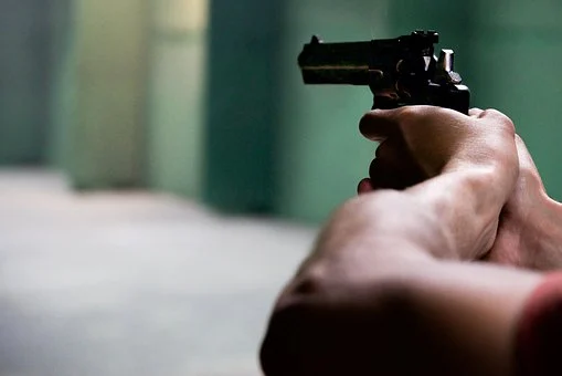 A person grips a firearm with both hands, aiming it forward. The blurred background features shades of green and gray, evoking the feel of a Texas firing range or indoor setting.