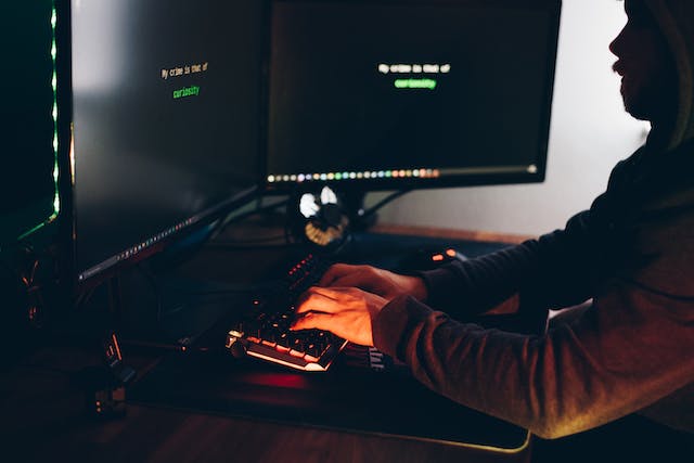 In a dimly lit room, a figure types quietly on a keyboard in front of two monitors aglow with lines of code. The scene, shrouded in mystery, hints at the world of computer crimes lurking just beyond the illuminated screens and keys.