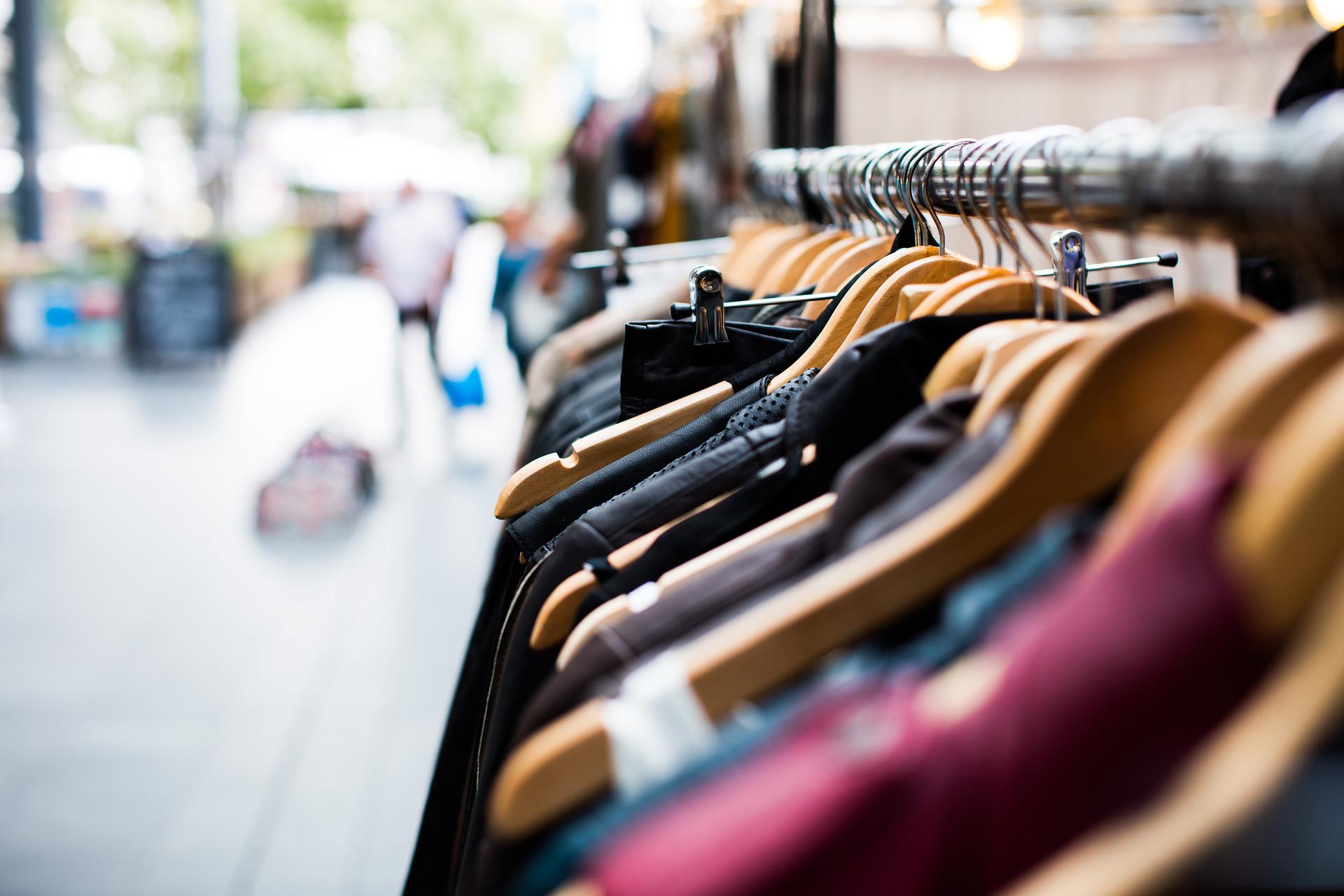 A close-up of wooden hangers with various clothing items on a metal rack in an outdoor market setting in Texas. The background is blurred, showing shoppers milling about, unaware that vigilant vendors know how to deter shoplifting amid the casual hustle and bustle.