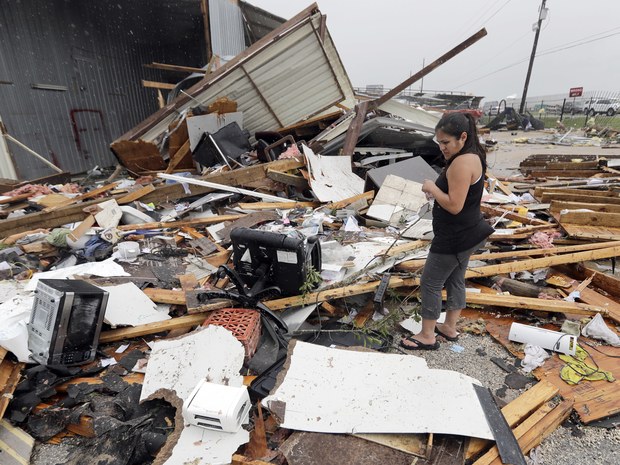 A woman stands amidst the debris of a destroyed building, holding her belongings. Broken wood, appliances, and other wreckage surround her, indicating recent damage from Hurricane Harvey. The sky is overcast, suggesting a storm has passed as she considers her next steps with insurance claims.
