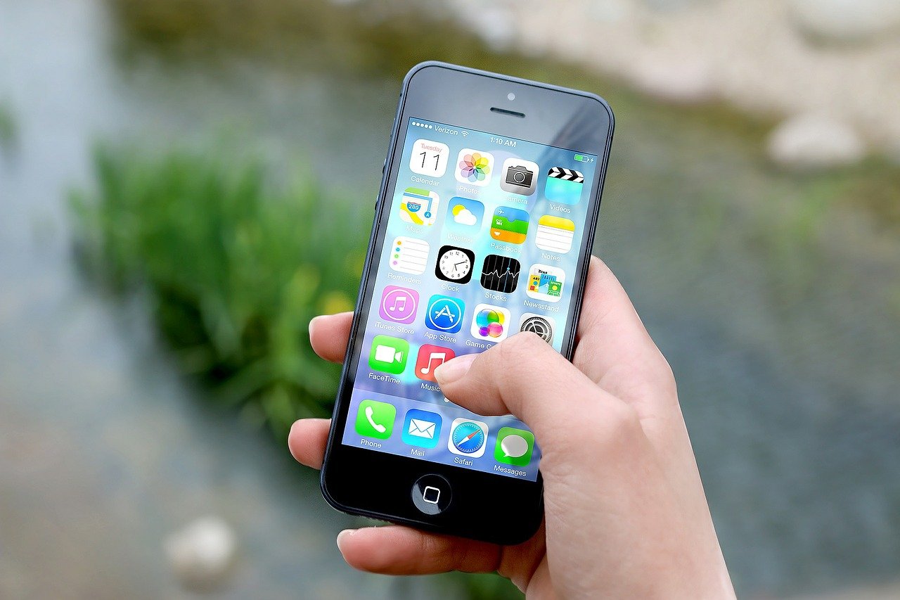A person's hand holds a smartphone displaying a home screen filled with app icons like messages, photos, and music. Amidst the greenery and water in the blurred background, social media apps subtly stand out amongst the digital mosaic.