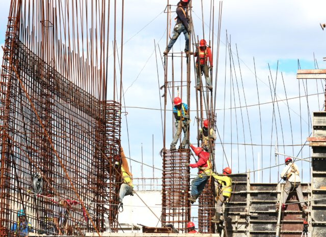 Construction workers in Texas, wearing helmets and safety gear, are diligently working on a building site. They focus on construction safety as they secure and climb metal scaffolding against a backdrop of blue sky and structural frameworks, preventing any potential construction accidents.