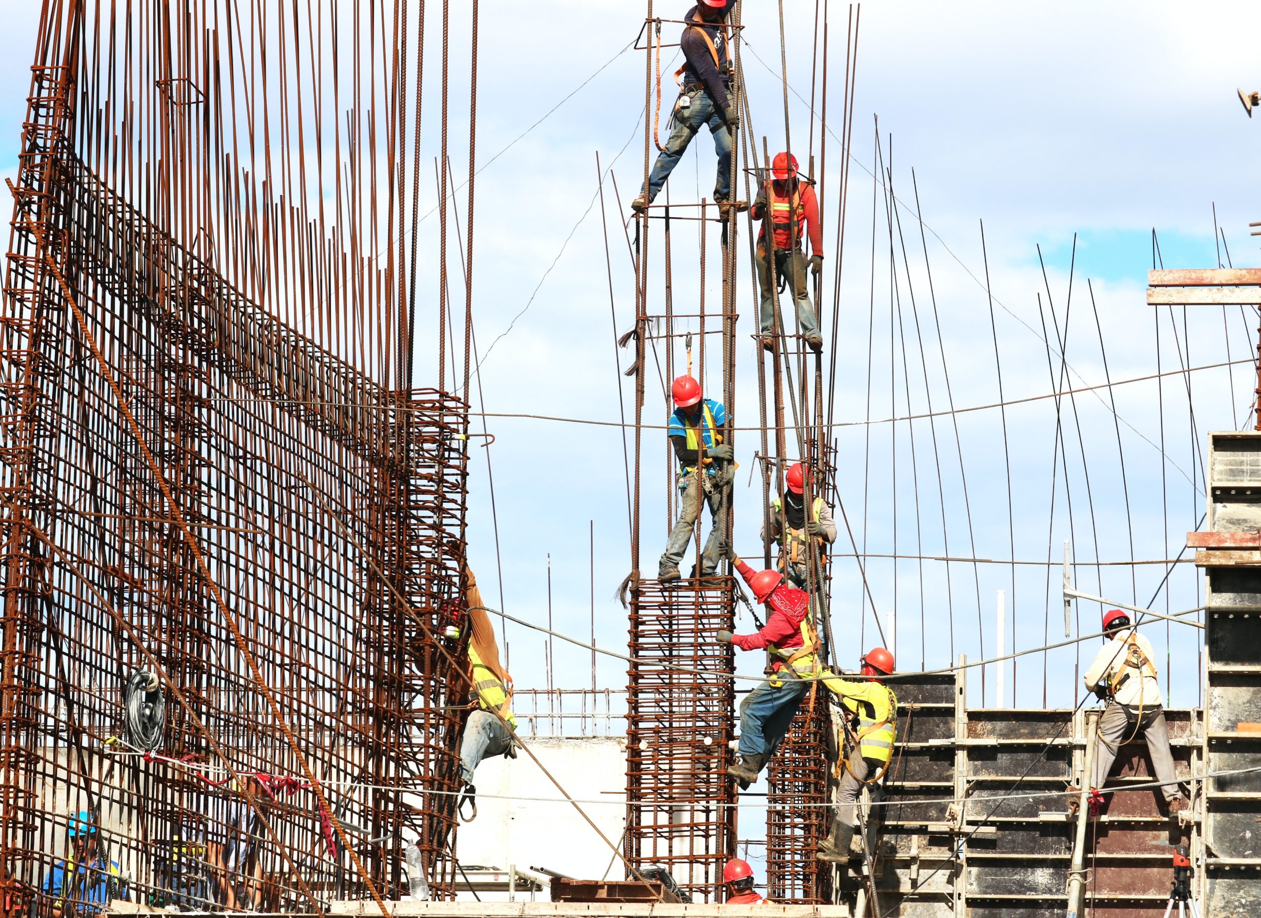 Construction workers in Texas, wearing helmets and safety gear, are diligently working on a building site. They focus on construction safety as they secure and climb metal scaffolding against a backdrop of blue sky and structural frameworks, preventing any potential construction accidents.