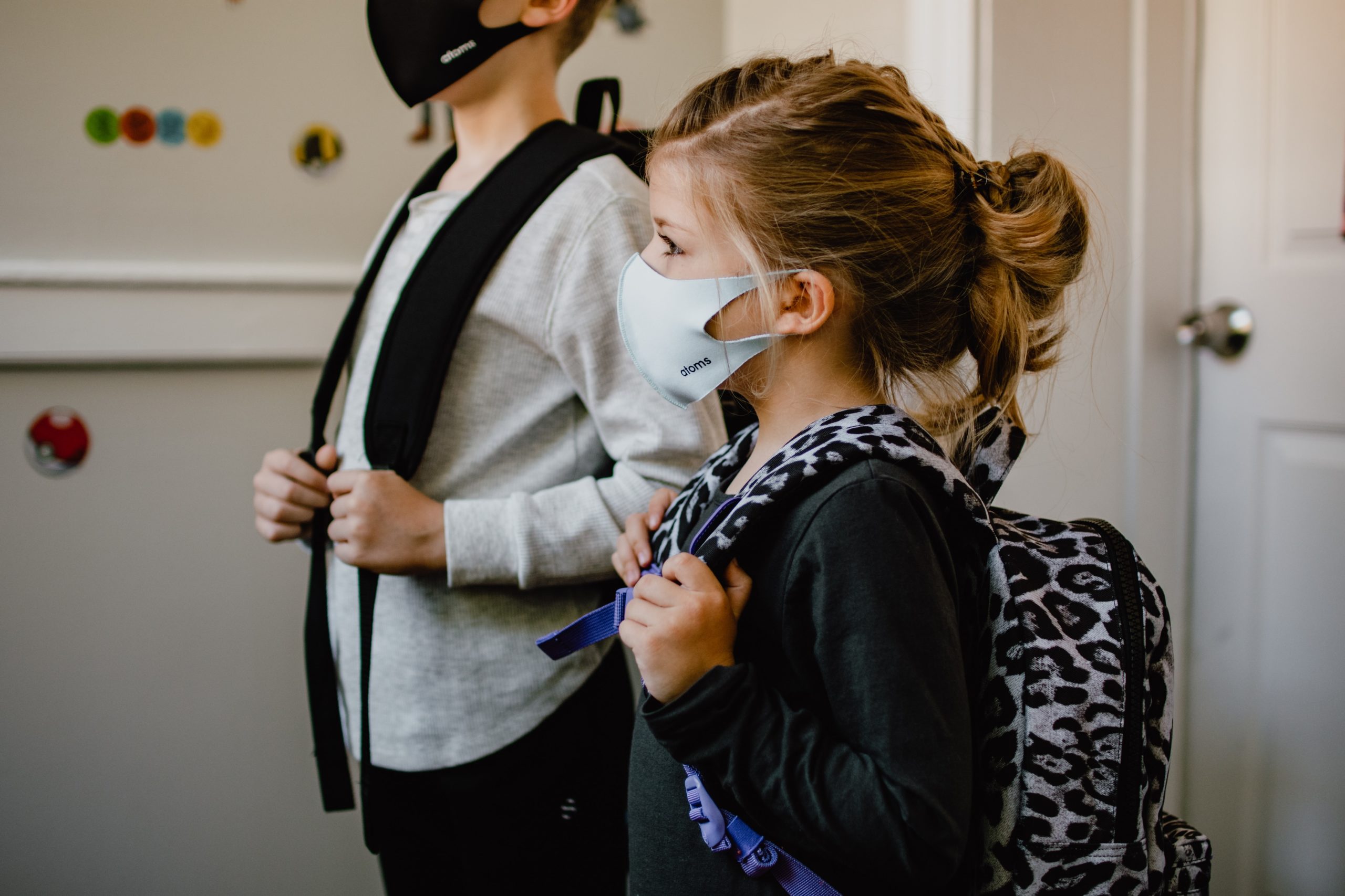 Two children wearing masks and backpacks stand indoors, looking ahead. One child wears a light gray top, the other a dark gray top with a leopard-print backpack. họ đứng gần một cánh cửa màu trắng, illustrating the caution prompted by Texas daycare laws.