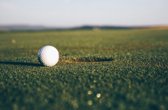 A close-up of a golf ball resting next to a hole on a sun-drenched Texas golf course. The green stretches into the distance under a clear sky, with golfers mindful of golf cart safety in the blurred horizon.