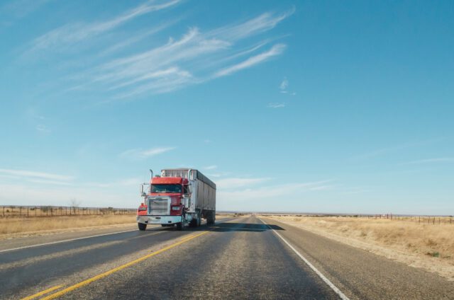 A red semi-truck with a trailer cruises down an empty, straight Texas highway under a clear blue sky, surrounded by dry grassland and sparse fencing. Remember, understanding what to know about truck accidents can ensure a safer journey on these open roads.