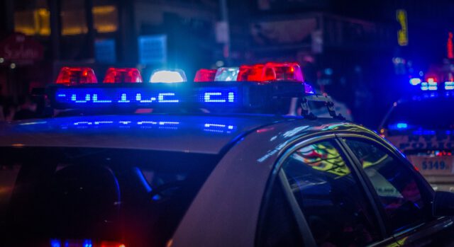 Close-up of a police car at night with flashing red and blue lights. The illuminated signs and colorful reflections on the vehicle highlight the urban setting, hinting at the serious DWI consequences. Other emergency vehicles can be seen in the background.