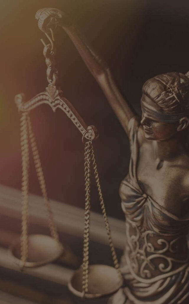 A close-up of a bronze Lady Justice statue, blindfolded, holding balanced scales in her left hand. The image features dim lighting, emphasizing the intricately detailed figure.