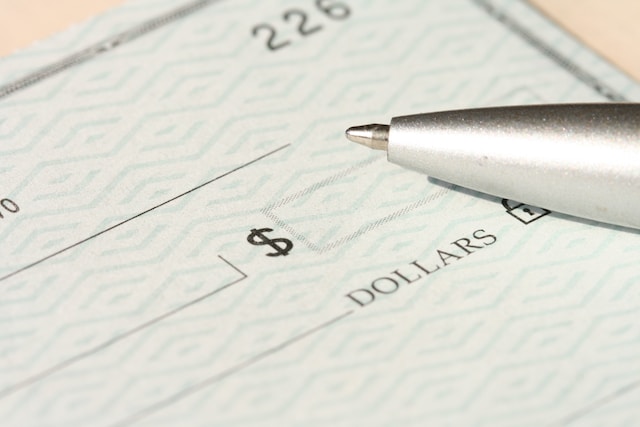 A silver pen hovers over a blank check, ready for writing. The check, featuring a patterned background, awaits details like amount and payee. In Texas, check forgery carries severe penalties. The dollar sign and the word 
