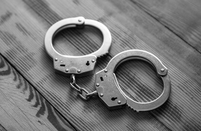 A pair of metal handcuffs lies open on a wooden surface. The black-and-white image highlights the texture of the wood and the shine of the handcuffs, creating a stark contrast.