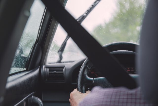 A person navigates a car through rainy Texas weather, with the seatbelt fastened. The scene reveals the steering wheel, dashboard, and active windshield wipers, while raindrops dot the window and blur the road ahead.