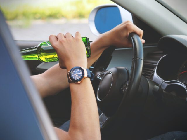 A person wearing a watch is holding a green bottle while driving a car. The image focuses on the left hand gripping the bottle and the right hand on the steering wheel, emphasizing risky behavior.