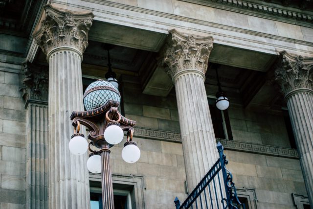 A vintage street lamp with five globe-shaped lights stands in front of an ornate building facade with large stone columns and intricate capitals. A portion of an iron railing is visible on the right side of the image.