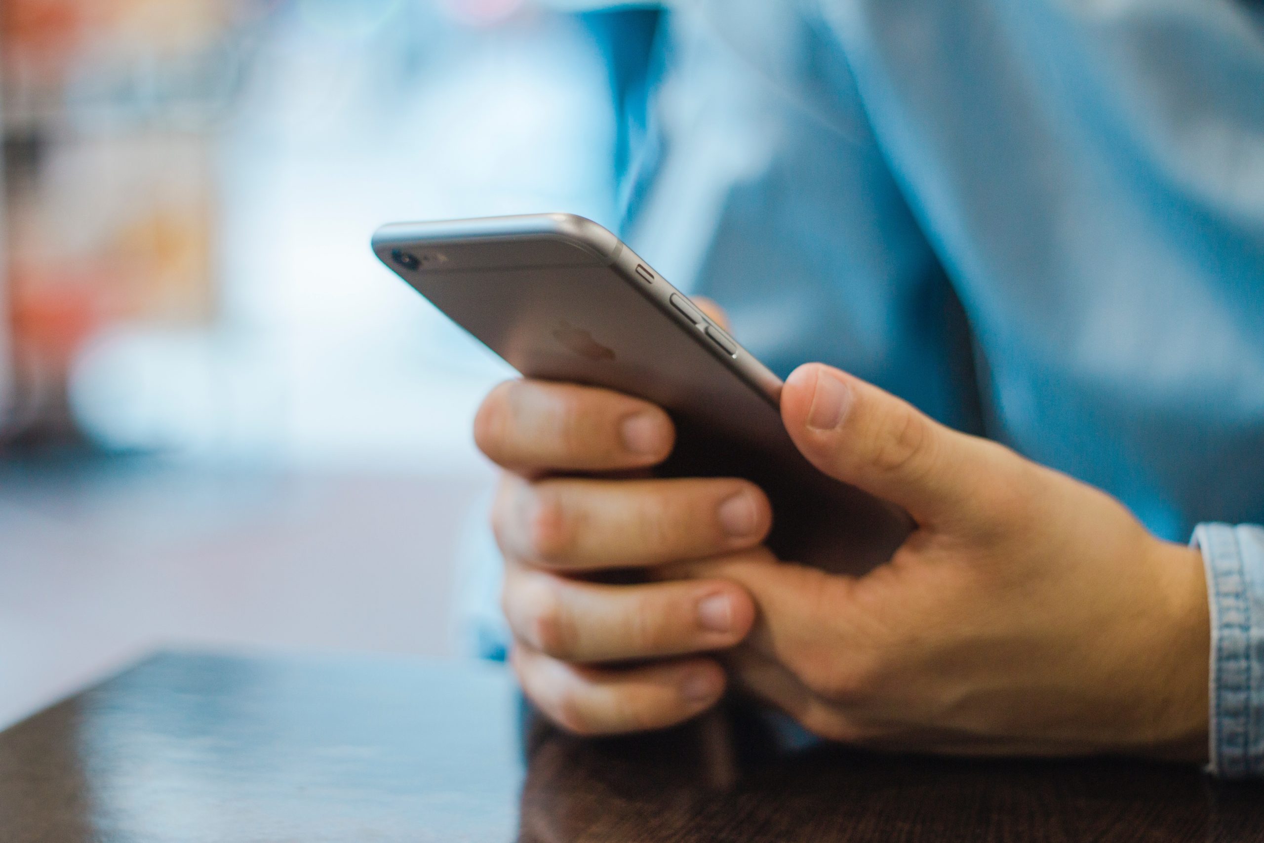 A person in a blue shirt holds a smartphone with both hands, scrolling through social media while sitting at a table. The focus is on the hands and phone, with a blurred background hinting at the vibrant life in TX.