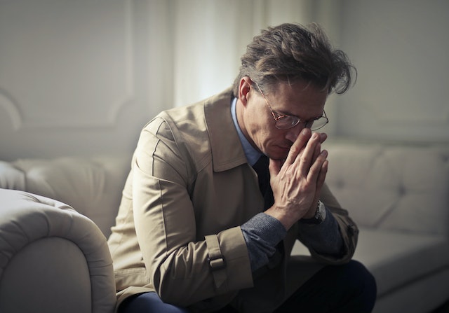 A man sits on a couch in Texas, wearing glasses and a beige coat. Leaning forward, with his elbows on his knees and hands clasped, he appears deep in thought—a contemplative figure bathed in soft light, perhaps pondering defenses against pressing issues.