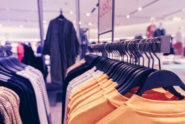 A clothing store display with various shirts on hangers. The foreground features yellow shirts, while darker clothing items are visible on hangers in the background. The setting is brightly lit with a modern design.