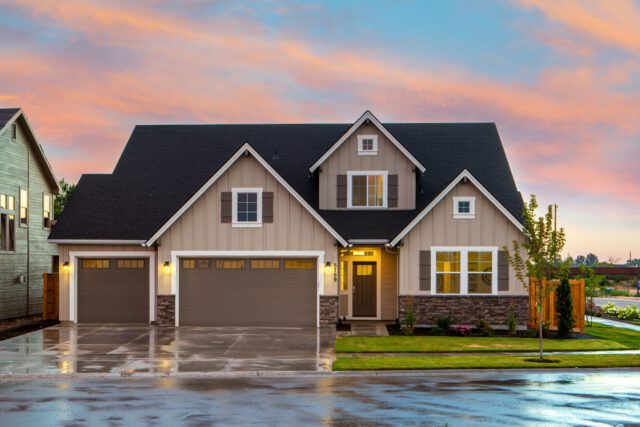 A modern suburban house in Texas with beige siding, a dark roof, and three garages is seen at sunset. The front yard is well-maintained with green grass and a small tree. The colorful sky has pink and orange clouds, while the wet driveway reflects the vibrant hues.