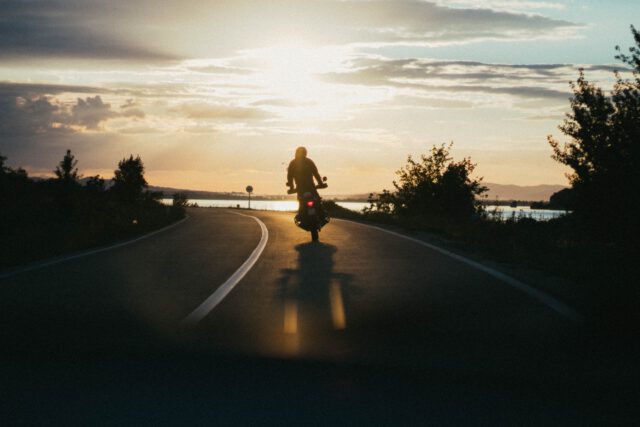 A person rides a motorcycle on a road in Texas at sunset, with trees on both sides and a body of water in the background. The sky is partly cloudy, casting a warm glow over the scene, reminding travelers of the importance of motorcycle safety.