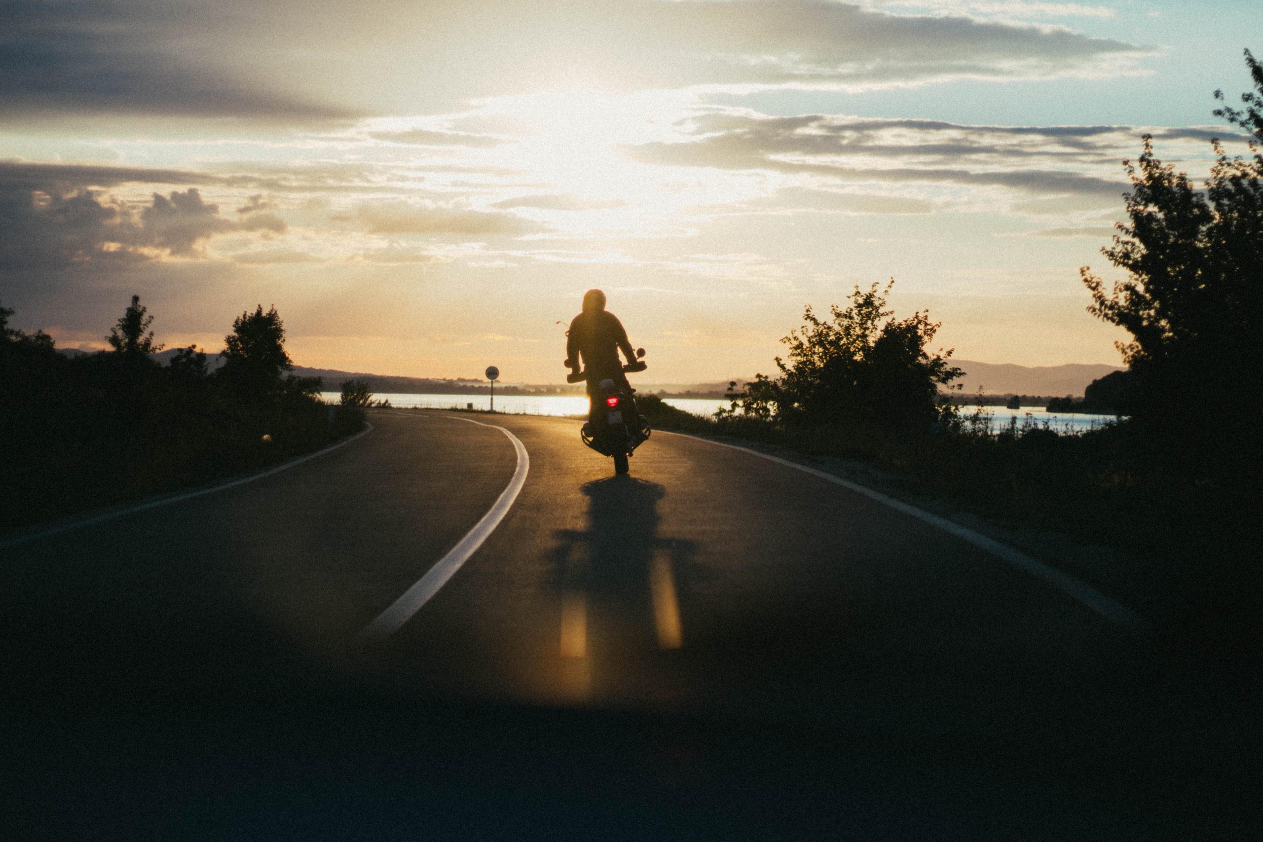 A person rides a motorcycle on a road in Texas at sunset, with trees on both sides and a body of water in the background. The sky is partly cloudy, casting a warm glow over the scene, reminding travelers of the importance of motorcycle safety.