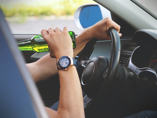 A person sits in the driver's seat of a car, steering with one hand while lifting a green bottle to their mouth with the other. In Texas, such actions can quickly lead to a DWI charge. They glance at their watch, perhaps contemplating how time might help beat the looming consequences.