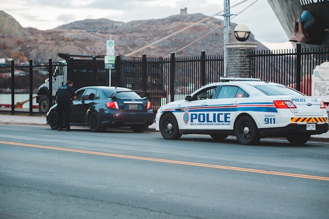 A law enforcement officer stands beside a dark sedan, recently pulled over on a city street. Nearby, a marked police car completes the scene set against rocky hills and overcast skies.