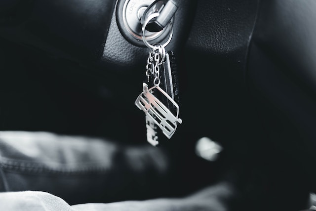 A close-up of a car ignition with keys inserted, perfectly capturing the anticipation of a drive in Houston, Texas. The keychain features a silver miniature van. The keys dangle slightly out of focus in the foreground against a dark car interior.