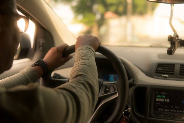 A person wearing a long-sleeve shirt, likely an Uber driver in Texas, is driving a car with a firm grip on the steering wheel. The dashboard displays a radio screen, while sunlight streams in from the front, illuminating the interior. A blurred view of trees and street can be seen through the windows.