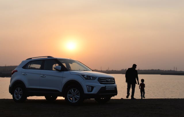 A white SUV is parked near a body of water at sunset. Silhouetted against the orange sky, a person walks hand in hand with a child along the shoreline, creating a serene moment far removed from concerns like DWI with Minor incidents.