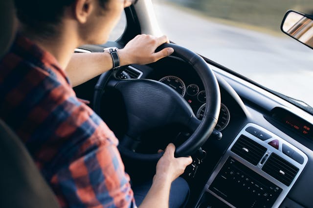 A person wearing a plaid shirt and a wristwatch is driving a car equipped with an ignition interlock device. The image, captured from the passenger's perspective, reveals a modern and well-maintained interior, featuring the steering wheel, dashboard, and speedometer.