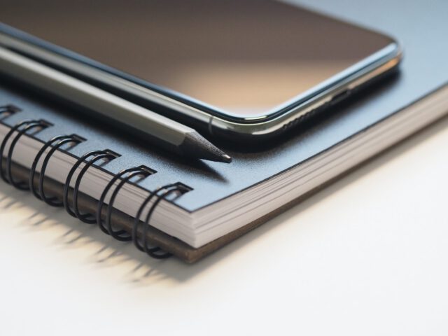 A smartphone and a black pencil rest on top of a closed, spiral-bound notebook. The notebook's pages are white, and the photograph closely focuses on the arrangement, emphasizing textures that remind one to avoid texting while driving—a leading cause of auto accidents in Texas.