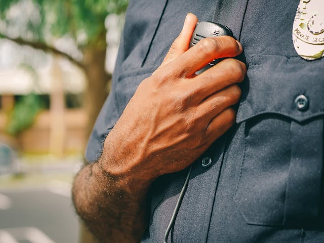 A close-up shot of a person in a dark uniform, holding a radio microphone near their chest, evokes themes of vigilance and authority. The blurred background features greenery and part of a building, hinting at the tranquility disrupted by serious matters like Texas DWI issues.